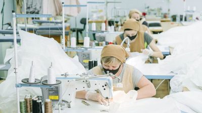 Women working on sewing machines in an industrial textile workshop, wearing protective masks and aprons.